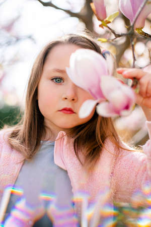 Portrait of a cute cheerful girl in a gray dress wearing rosa jacket staying in spring in a park under a flowering pink magnolia tree.の写真素材