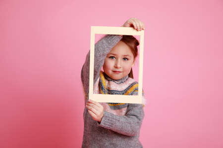 Little girl in warm dress looking through frame on rosa bachground.の写真素材