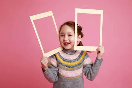 Portrait of a little smiling girl holding blank frame for mock-up on a pink background.の写真素材