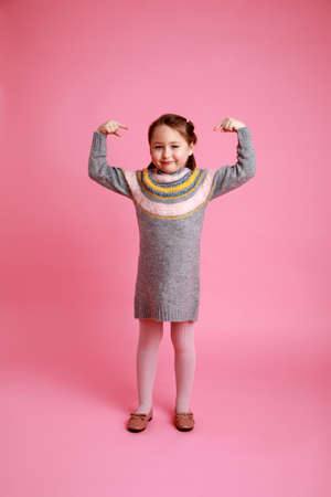 Portrait of little funny smiling girl showing to themself in warm dress on pink background.の写真素材