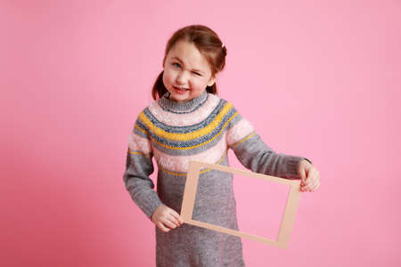 Portrait of a little smiling girl holding blank frame for mock-up on a pink background.の写真素材