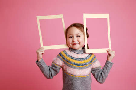 Portrait of a little smiling girl holding blank frame for mock-up on a pink background.の写真素材