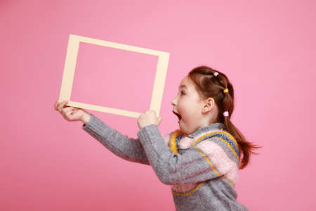 Portrait of a little smiling girl holding blank frame for mock-up on a pink background.の写真素材