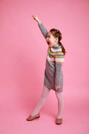 Portrait of little funny smiling girl showing to themself in warm dress on pink background.の写真素材