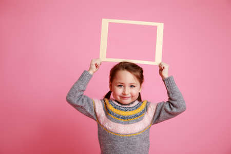 Portrait of a little smiling girl holding blank frame for mock-up on a pink background.の写真素材