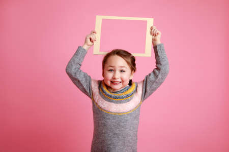 Portrait of a little smiling girl holding blank frame for mock-up on a pink background.の写真素材
