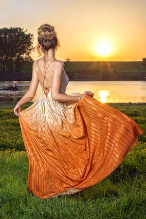 Woman in a long glamorous dress at sunset. Beautiful landscape view, shot from the backの写真素材