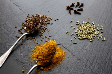 Fennel seeds, cloves, caraway seeds and curry powder on a slate boardの写真素材