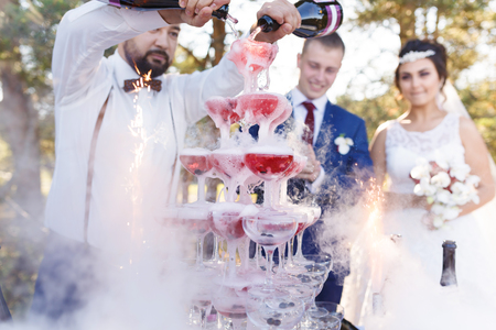 Bartender with newlyweds are pouring vine in the pyramid of glasses at the open air wedding banquetの写真素材
