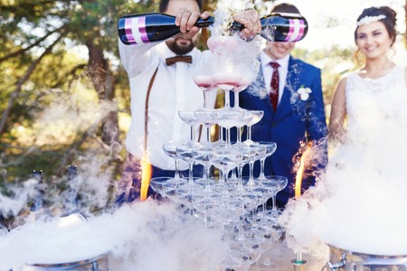 Bartender with newlyweds are pouring vine in the pyramid of glasses at the open air wedding banquetの写真素材
