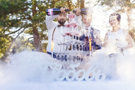 Bartender with newlyweds are pouring vine in the pyramid of glasses at the open air wedding banquetの写真素材