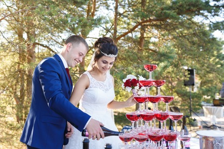 Newlyweds are pouring vine in the pyramid of glasses at the open air wedding banquetの写真素材