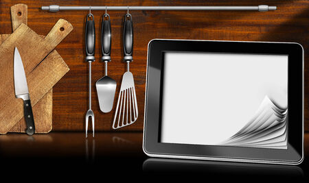 Black tablet computer with blank pages in a kitchen, on wooden wall with kitchen utensils. Template for recipes or food menuの写真素材