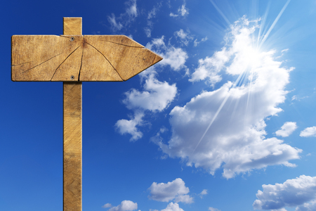 Wooden directional sign with one empty arrow on a blue sky with clouds and sun raysの写真素材