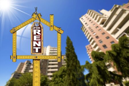 Yellow wooden meter ruler in the shape of house and label with text for rent. For rent real estate sign with tall and blurred buildings in the backgroundの写真素材