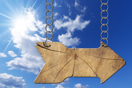 Wooden directional sign with one empty arrow hanging with metal chain on blue sky with clouds and sun raysの写真素材