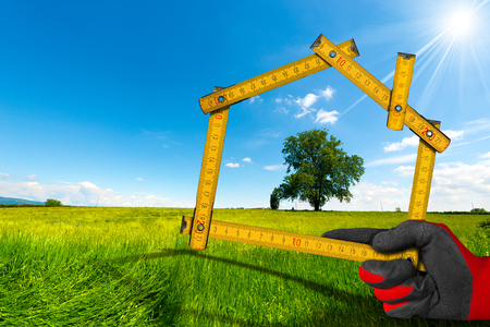 Hand with work glove holding a wooden meter ruler in the shape of house, in the countryside with green tree. Concept of ecological house projectの写真素材