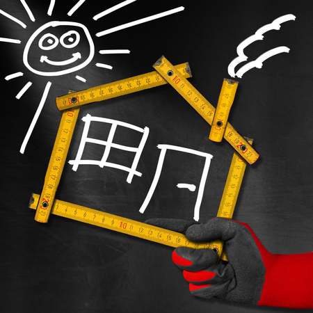 Hand holding a wooden meter ruler in the shape of house with sun, door, window and smoke from the chimney. On a blackboard. Concept of house projectの写真素材