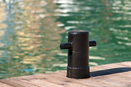 Detail of old and black mooring bollard on the wooden pier with water in the background