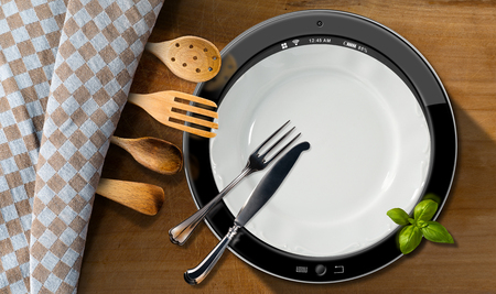 Round tablet computer in a kitchen with silver cutlery and wooden kitchen utensils on a wooden table with checkered tableclothの写真素材