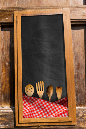 Empty blackboard with red and white checkered tablecloth and four wooden kitchen utensils. Template for a food menuの写真素材