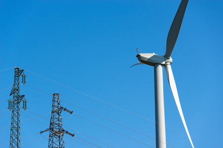 Detail of a white wind turbine with a power line (two high voltage towers) on a clear blue sky