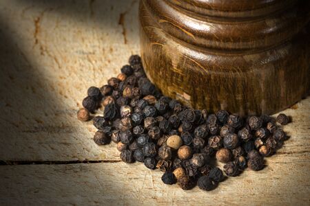 Macro photo of peppercorns and an old wooden pepper mill on wooden cutting board with dark shadowsの写真素材
