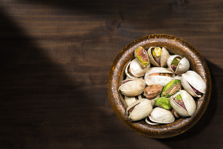 Roasted and salted pistachios in a wooden bowl on a brown table with dark shadows - Macro photographyの写真素材