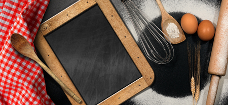 Baking background with empty blackboard on a black table with  Rolling pin, whisk, eggs, spoons, ears of wheat and a red and white checkered tableclothの写真素材