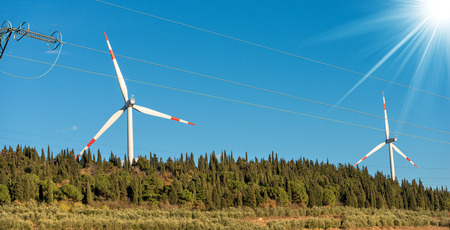 Two red and white wind turbines with a power line on a clear blue sky with sun rays. Renewable...
