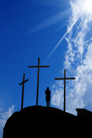 Silhouette of three crosses on the hill and a boy praying on a blue sky with clouds and sun raysの写真素材