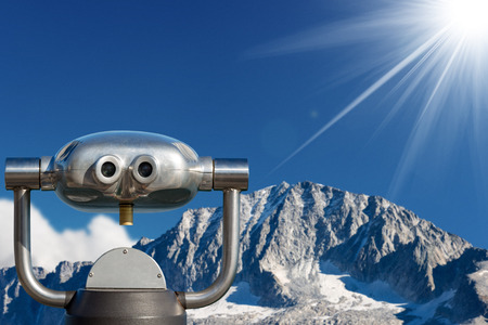 Coin operated electronic binoculars for tourists on a blurred landscape, mountain peak with glacier and blue sky with sun raysの写真素材