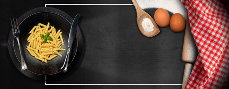Wooden plate with Italian Pasta on a blackboard with copy space, kitchen utensils, flour, rolling pin, eggs, checkered tablecloth, fork and knifeの写真素材