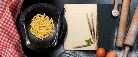 Italian pasta called Penne and an empty recipe book on a table with kitchen utensils, flour, rolling...