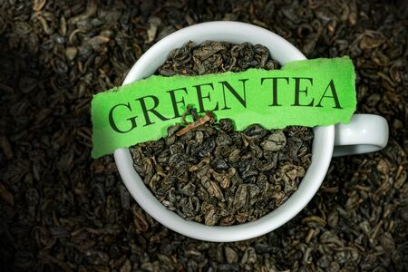 Close-up of dried leaves of green gunpowder tea in a white cup and piece of paper with text Green Tea, with many rolled up leaves on the backgroundの写真素材