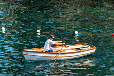 GULF OF LA SPEZIA, LIGURIA, ITALY - JULY 21th, 2019: A man on a white small wooden rowboat in the green Mediterranean sea in front of the ancient Tellaro village, Liguria, Italy, Europeのeditorial素材