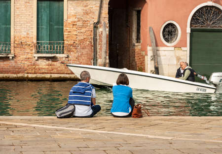 A couple of tourists sitting on the edge of a canal while a motorboat sails in front of them. Venice, UNESCO world heritage site, Veneto, Italy, Europeのeditorial素材