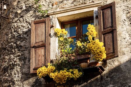 Close-up of an old window with wooden shutters and yellow flowers in the medieval village of Canale di Tenno or Villa Canale, Italian Alps, Trento province, Trentino-Alto Adige, Italy, Europeの写真素材