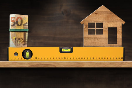 Construction spirit level with above a roll of Euro banknotes and a small wooden model house. Above...