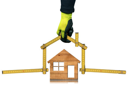 Closeup of a hand with work glove holding a folding ruler in the shape of a house above a wooden model house. Isolated on white background. Construction industry concept.の写真素材