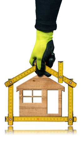Gloved hand holding a folding ruler in the shape of a house with a small wooden model house inside. Isolated on white background with reflections. Construction industry concept.の写真素材