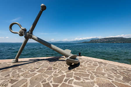 Large anchor in front of the port of Bardolino village, tourist resort on the coast of Lake Garda (Lago di Garda). On the horizon the headland called Punta San Vigilio. Verona, Veneto, Italy, Europe.の写真素材