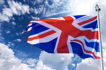 Close-up of a national UK flag with flagpole, blowing in the wind on a blue sky with clouds and copy...