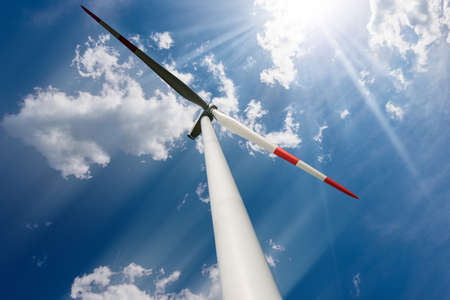 Red and white wind turbine against a blue sky with clouds, sun rays and copy space. Renewable energy...