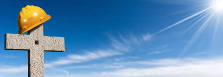 Orange hard hat or safety helmet on top of a stone cross on a clear blue sky with clouds and copy...