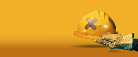 Manual worker with protective work glove holding a yellow safety helmet with a cross plaster (adhesive bandage), on a yellow and orange background with copy space and reflections. Workplace security.の写真素材