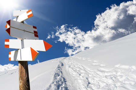 Blank directional trail signs in a beautiful winter mountain landscape with powder snow and footprints against a clear blue sky and sunbeams. Italian Alps, Italy, Europe.の写真素材