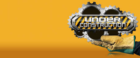 Gloved hand holding an under construction sign with metal cogwheels (gears), on orange and yellow...