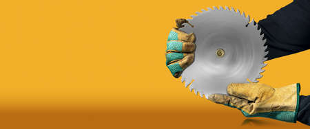 Carpenter with protective work gloves holding a metal circular saw blade, on orange and yellow background with copy space and reflections.の写真素材
