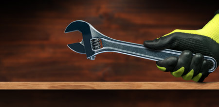 Manual worker with protective black and green work gloves holding a stainless steel adjustable wrench, on a wooden workbench with copy space.の写真素材